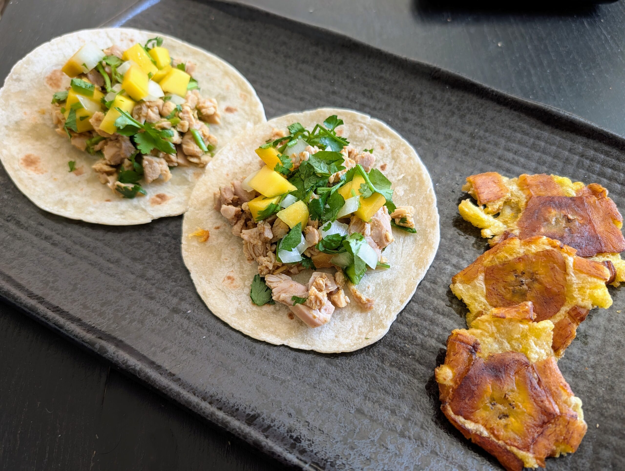 Two tacos with diced mango on a plate next to fried plantain slices.